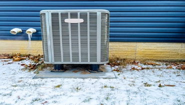 An electric heat pump and ac unit is outside a blue vinyl sided house in the winter time.