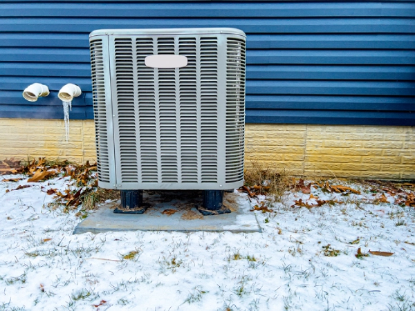 an-electric-heat-pump-and-ac-unit-is-outside-a-blue-vinyl-sided-house-in-the-winter-time