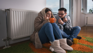A man and woman sit next to their radiator while wrapped in blankets, indicating that their home’s heating system is broken. | Aire Serv of North Central Arizona.