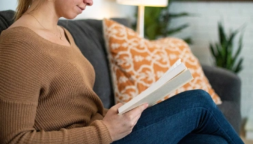 Woman reading on a gray couch next to an orange and white throw pillow with two plants in the background.