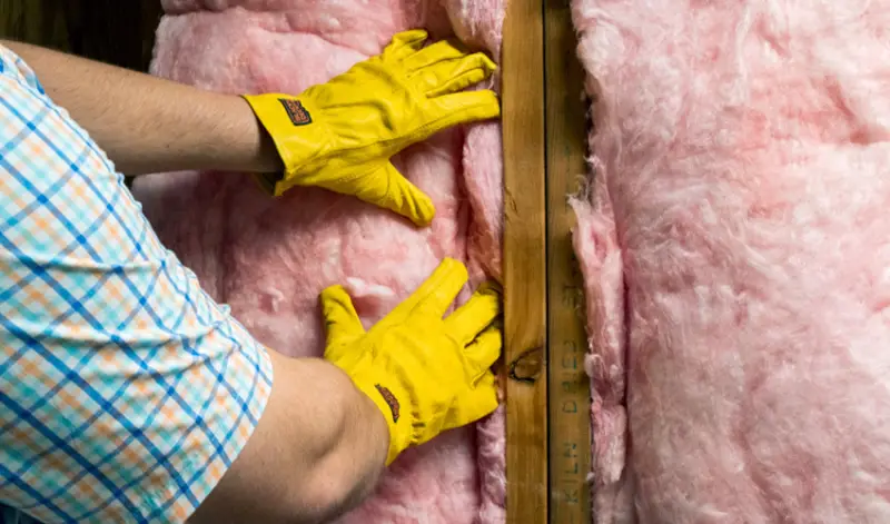 Yellow-gloved arms manipulating pink insulation in a wall