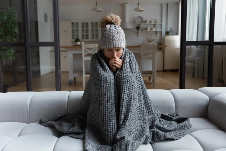  A woman wrapped in a blanket on her couch because her home&rsquo;s heater is broken.